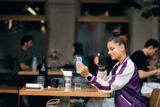 Young woman sitting outdoors on terrace in cafe at the table drinking beverage tea or coffee holding in hand smartphone