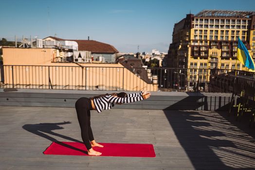 Young sporty happy woman in sportswear doing stretching exercises on yoga mat on house roof in early morning