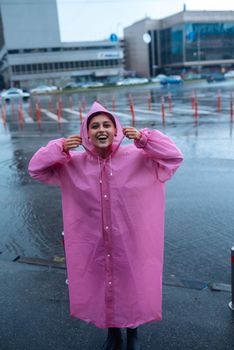 Young smiling woman with a pink raincoat on the street while enjoying a walk through the city on a rainy day.
