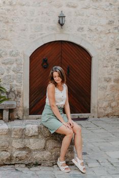 Girl Tourist Resting in the Ancient Narrow Street On A Beautiful Summer Day In MEDITERRANEAN MEDIEVAL CITY, OLD TOWN KOTOR, MONTENEGRO. Young Beautiful Cheerful Woman Walking On Old Street