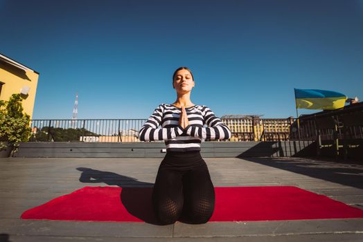 Young sporty happy woman in sportswear doing stretching exercises on yoga mat on house roof in early morning