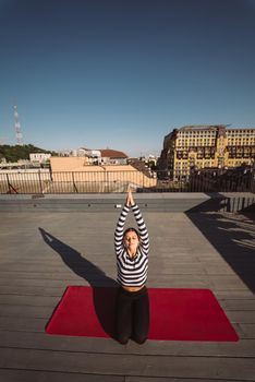 Young sporty happy woman in sportswear doing stretching exercises on yoga mat on house roof in early morning