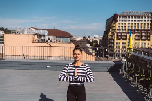 Young sporty happy woman in sportswear doing stretching exercises on yoga mat on house roof in early morning