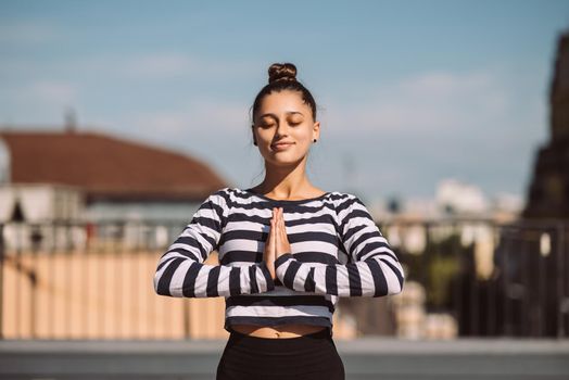 Young sporty happy woman in sportswear doing stretching exercises on yoga mat on house roof in early morning
