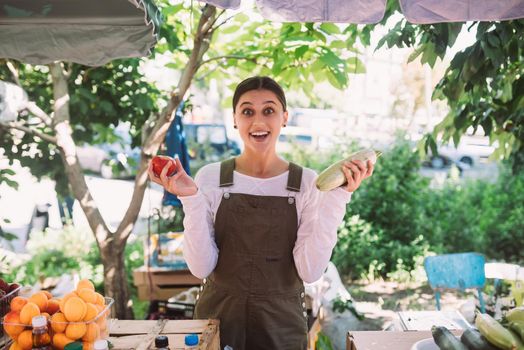 Young positive salesgirl at job, selling sells home-grown vegetables and fruits in vegetable shop