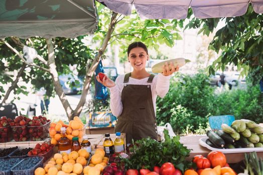 Young positive salesgirl at job, selling sells home-grown vegetables and fruits in vegetable shop