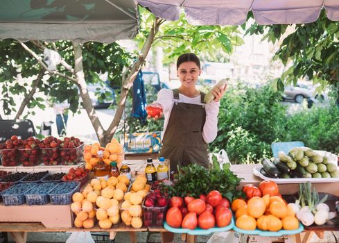 Young positive salesgirl at job, selling sells home-grown vegetables and fruits in vegetable shop
