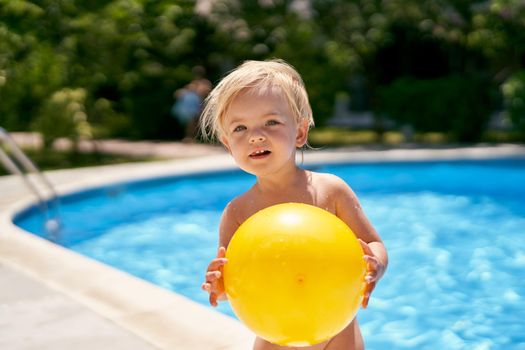 Small curious baby with a yellow ball stands by the pool. High quality photo