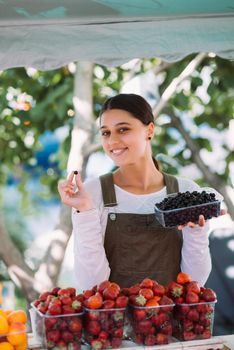 Young positive salesgirl at job, selling sells home-grown vegetables and fruits in vegetable shop
