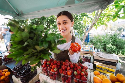 Young positive salesgirl at job, selling sells home-grown vegetables and fruits in vegetable shop