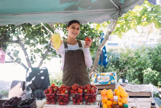 Young positive salesgirl at job, selling sells home-grown vegetables and fruits in vegetable shop