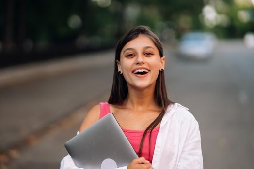 A young woman holding a laptop looks at the camera on the street