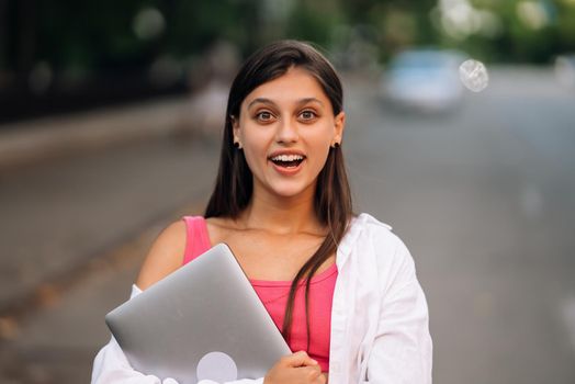 A young woman holding a laptop looks at the camera on the street