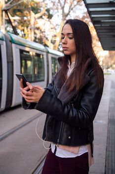 vertical photo of a woman consulting her telephone at the streetcar stop, concept of technology and public transportation