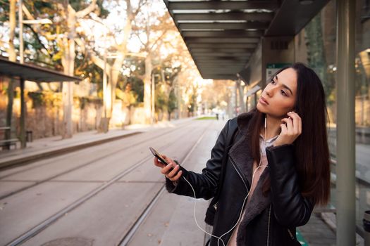 woman putting on her telephone earphones at the streetcar stop, concept of technology and communication, copyspace for text