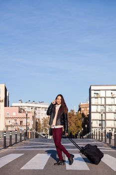 vertical photo of a young woman pulling suitcase talking by phone while crossing the street at a pedestrian crosswalk, concept of travel and urban lifestyle, copyspace for text