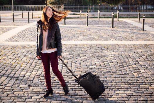 happy young woman with trolley suitcase walking down a cobblestone street, concept of travel and urban lifestyle, copyspace for text