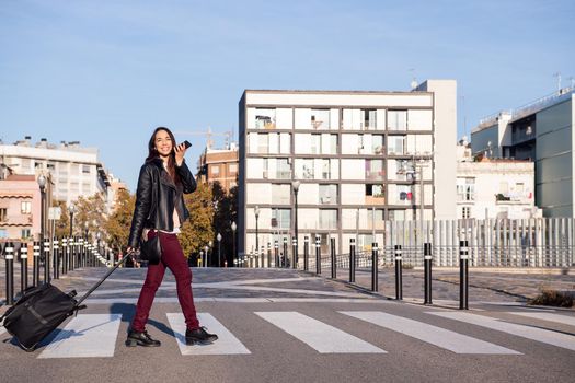 smiling woman pulling suitcase recording a voice message with phone while crossing the street at a pedestrian crosswalk, concept of travel and urban lifestyle, copyspace for text
