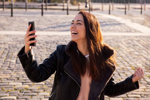 joyful young woman laughing and celebrating looking the phone on a cobblestone street, concept of technology and urban lifestyle, copyspace for text