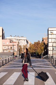 vertical photo of a woman pulling suitcase recording a voice message with phone while crossing the street at a pedestrian crosswalk, concept of travel and urban lifestyle, copyspace for text