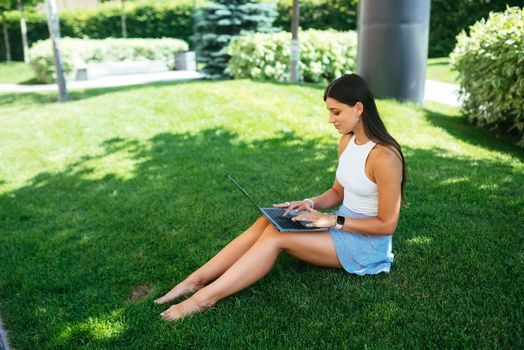 The concept of remote work. A young girl works with a laptop in the fresh air in the park, sitting on the lawn.