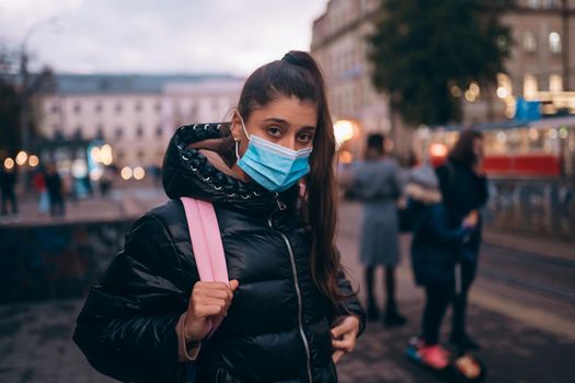 Portrait of beautiful young student woman in medical mask posing outdoors with a backpack