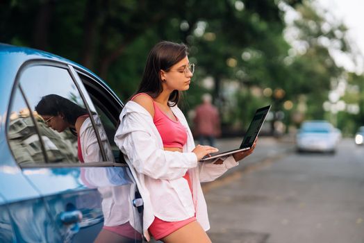 A young woman uses a laptop near a car in city center outdoors