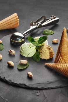 Close-up shot of a fresh pistachio and chocolate ice cream decorated with fresh mint, waffle cones and scattered nuts, served on a stone slate over a black background. A metal scoop is laying nearby. Top view.