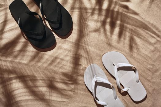Summer concept with a shadow of a tropical palm tree leaves, copyspace. White and black thongs are on a perfect beach sand. Summertime lifestyle, objects in flat lay, top view arrangement.