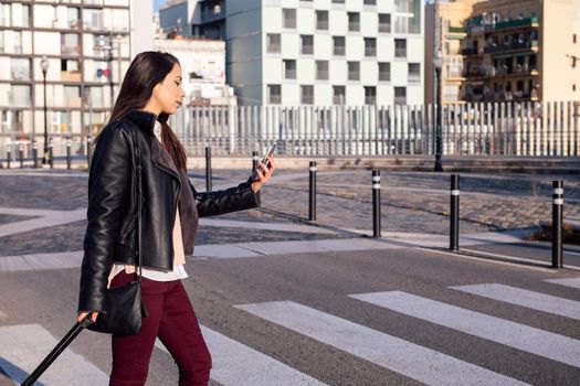 woman with trolley suitcase consulting phone while crossing the street at a pedestrian crosswalk, concept of travel and urban lifestyle, copyspace for text