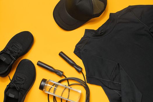 Close-up shot of a black gym accessories, such as sport shoes, t-shirt, transparent bottle, baseball cap and skipping rope on a yellow background. Top view, flat lay. Fitness and healthy lifestyle concept.