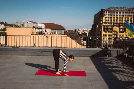 Young sporty happy woman in sportswear doing stretching exercises on yoga mat on house roof in early morning