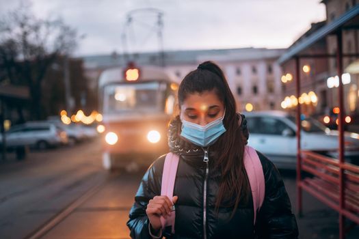Portrait of beautiful young student woman in medical mask posing outdoors with a backpack