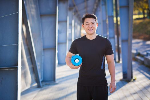 Active handsome male yogi walks in a stadium strolling before or after a workout. Young happy asian athletic man athlete goes to yoga class with a fitness mat in his hands Outdoors Healthy lifestyle