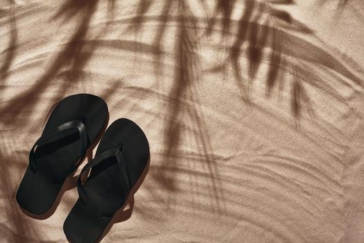 Summer concept with a shadow of a tropical palm tree leaves, copyspace. Black thongs are on a white beach sand. Summertime lifestyle, objects in flat lay, top view arrangement.