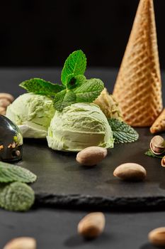 Close-up shot of an enjoyable pistachio ice cream decorated with fresh mint and scattered nuts, served on a stone slate over a black background. Side view.