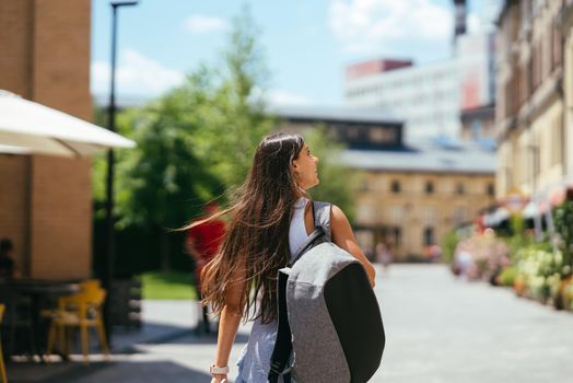 Urban young hipster woman with stylish backpack walks around the city near a building in the spring day. Pretty girl travels.