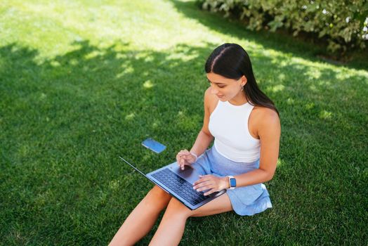 The concept of remote work. A young girl works with a laptop in the fresh air in the park, sitting on the lawn.