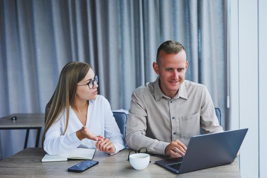 Businessmen are discussing while working with laptop in the office. Focused business people cooperating in a modern workspace. Two young businessmen are sitting together at a table.