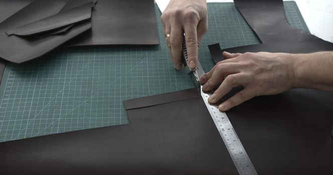 Close up of leather craftsman working with natural leather by knife and ruler by cutting a leather to a peaces. Handmade master at work in local workshop. Leather wallet stitch