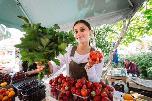 Young positive salesgirl at job, selling sells home-grown vegetables and fruits in vegetable shop
