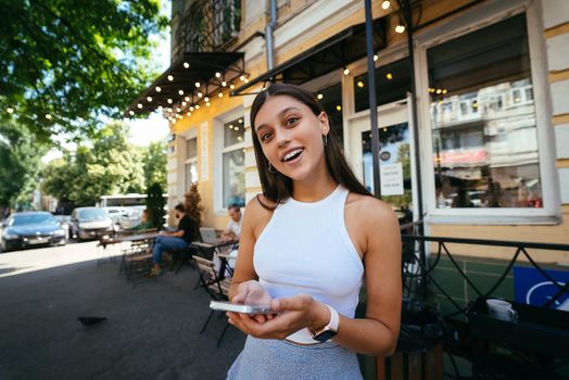 Smiling woman holding phone outdoors in cafe. A beautiful model looks at the camera