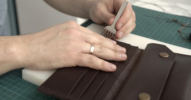 Mens hand holding a hammer and hole punch and makes a holes for seawing a leather wallet in his workshop. Working process with a brown natural leather. Craftsman holding a crafting tools