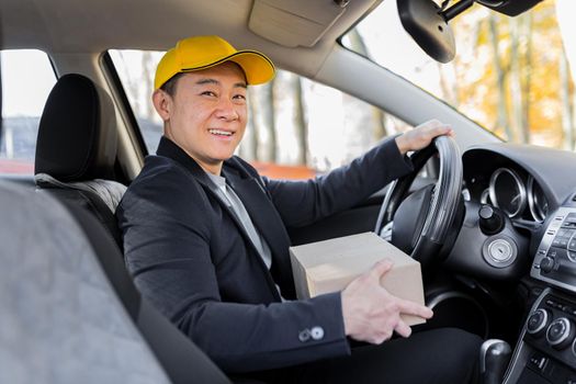 Male courier asian looks into the camera and smiles holding a parcel sitting in the car