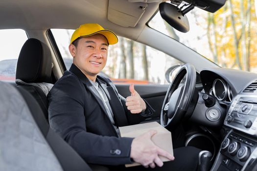 Male courier asian looks into the camera and smiles holding a parcel sitting in the car