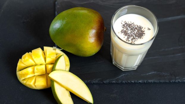 Milk drink on black background with mango. A classic mango milkshake - Lassi. A traditional drink in India from the heat with chia seeds. Turmeric lassie or mango lassi on black background with copy space. Indian traditional cold drink with mango. Freshness lassi made of yogurt, water, spices, fruits and ice. Top view