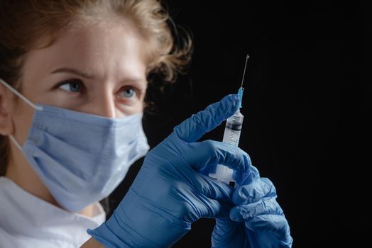 A female doctor wearing a protective mask and gloves holds a syringe with a vaccine or injection against a black background. A female nurse prepares to give an injection against a dark background. Woman with syringe in hand alone on black background. A drop of liquid on the syringe.