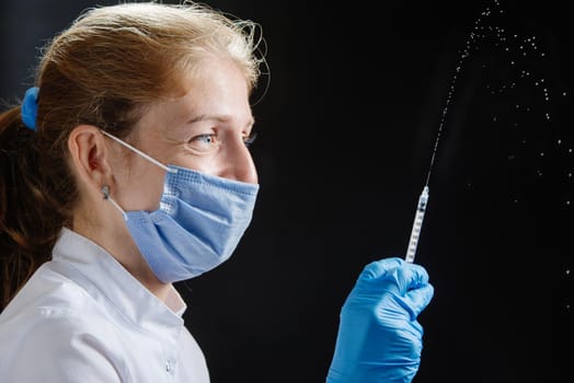 the doctor sprays from a syringe against a black background. The doctor smiles under his protective mask, the drug from the syringe pours into the air. A woman doctor wearing a medical mask, gloves and squirts liquid from a large syringe on a blue background.