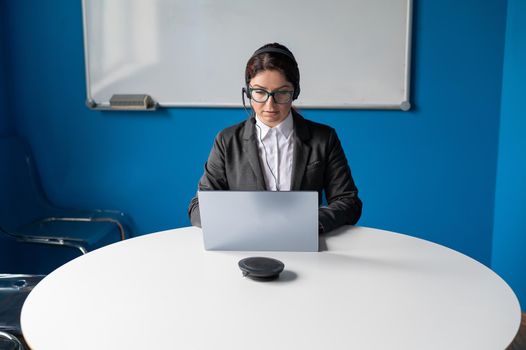 Serious woman wearing a headset is having an online conversation on a laptop in a conference room. Female boss communicates subordinates by video call. An employee is watching a business webinar