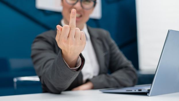 Angry business woman in a suit shows a middle finger while sitting at a desk. Annoyed female office worker showing a fuck you gesture. Professional burnout